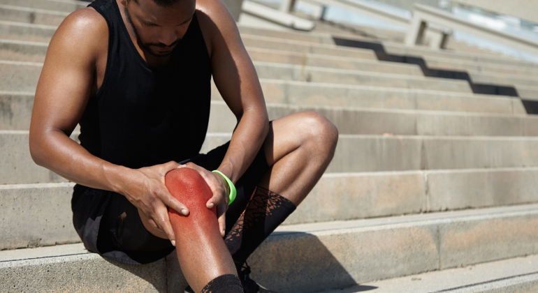 Man sitting on outdoor steps, holding his sore knee in pain. He is wearing a black tank top and shorts.