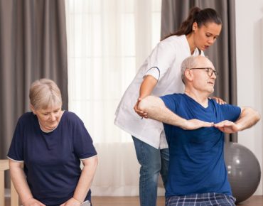 A physiotherapist assisting an elderly man with arm exercises while an elderly woman performs seated exercises in a home setting, featuring comfortable surroundings and fitness equipment in the background.
