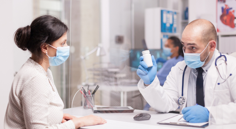 Doctor wearing a mask and gloves showing a medication bottle to a masked patient across a desk in a medical office.