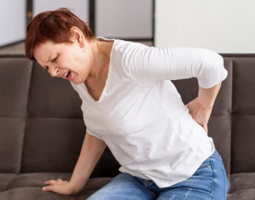 Woman sitting on a couch, grimacing in pain while holding her lower back.