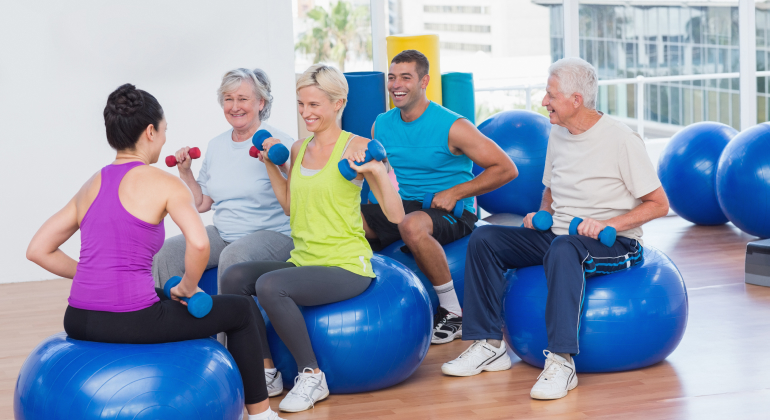 A group of people of various ages participating in a fitness class, sitting on large exercise balls and holding small dumbbells. They are smiling and engaged in their workout, which takes place in a bright and spacious fitness studio with large windows offering a view of the city. The atmosphere is lively and encouraging, promoting health and wellness through group exercise.