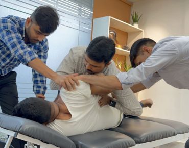 A team of three physiotherapists working together to perform advanced manual therapy on a patient lying on a treatment table in a modern clinic. The therapists are focused on adjusting the patient's upper body and hip area to improve mobility and relieve pain.