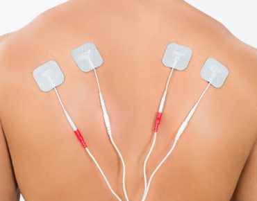 Close-up of a person's back with four adhesive electrode pads placed for electrical stimulation therapy. The pads are connected to wires, used to relieve muscle pain and tension. The background is plain and white, focusing attention on the treatment area.