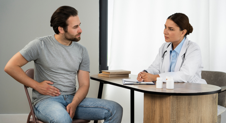 Doctor consulting with a male patient who is holding his stomach, indicating discomfort. They are seated across a desk in a clinical setting.