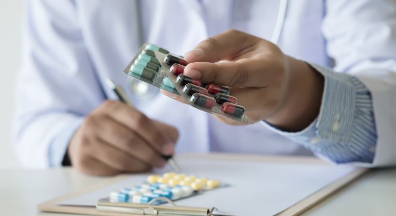 Close-up of a doctor holding a blister pack of pills while writing on a clipboard in a medical office.