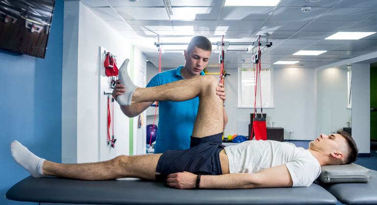 A physiotherapist helping a young man with a leg exercise in a modern clinic. The patient is lying on his back on a treatment table, with one leg bent and lifted while the therapist supports and guides the movement. The clinic is equipped with various rehabilitation equipment and has a professional and clean environment.