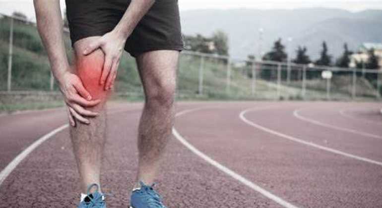 A person standing on a running track, clutching their knee with both hands. The knee is highlighted with a red area to indicate pain or injury. The person is wearing athletic shorts and blue running shoes, suggesting they might be experiencing sports-related knee pain. The background includes a blurred view of the track, grassy area, and distant mountains.