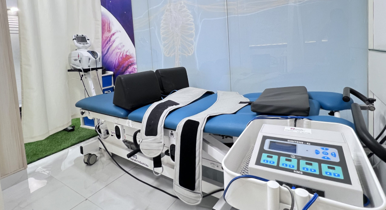 A physiotherapy treatment room equipped with modern rehabilitation equipment, including a treatment table with straps and cushions, an electronic therapy machine, and other medical devices. The room has a clean and professional setup with a background featuring anatomical illustrations.