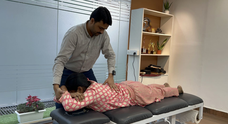 A physiotherapist performing a back massage on a woman lying face down on a treatment table in a modern clinic. The therapist is focused on providing relief and improving the patient's back health. The clinic is well-lit with a glass partition and shelves displaying various awards and plants in the background.