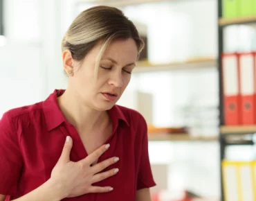 Woman in a red shirt clutching her chest in discomfort while standing in an office setting.