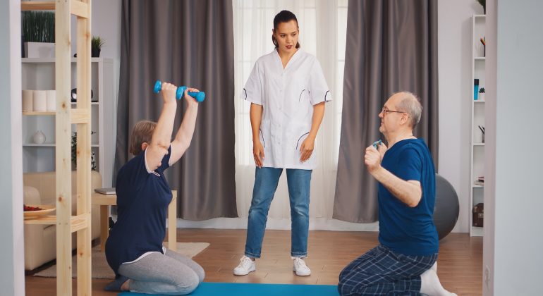 A physiotherapist overseeing an elderly man and woman performing exercises on a mat in a home setting. The woman lifts small dumbbells while kneeling, and the man engages in an arm exercise. The environment features home furnishings and fitness equipment in the background.