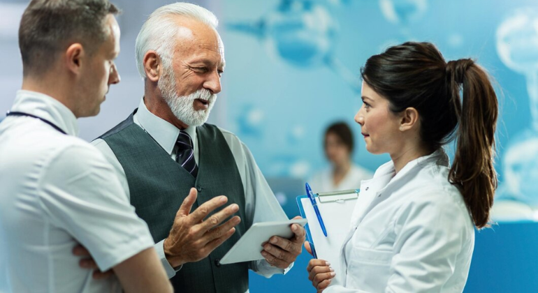 The image shows a group of three professionals engaged in a discussion, likely within a medical or healthcare setting. An older man with white hair and a beard, dressed in a suit vest and tie, appears to be leading the conversation, holding a tablet. He is speaking with a younger woman in a lab coat who holds a clipboard, suggesting she might be a doctor or healthcare professional. Another man, also in a white coat, listens attentively. The background features a modern, clean environment with a blue color scheme, possibly indicative of a medical or research facility.