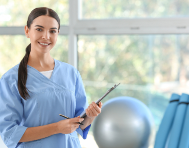 Smiling female physiotherapist in a light blue uniform holding a clipboard, standing in a bright, well-equipped therapy room with exercise equipment in the background.