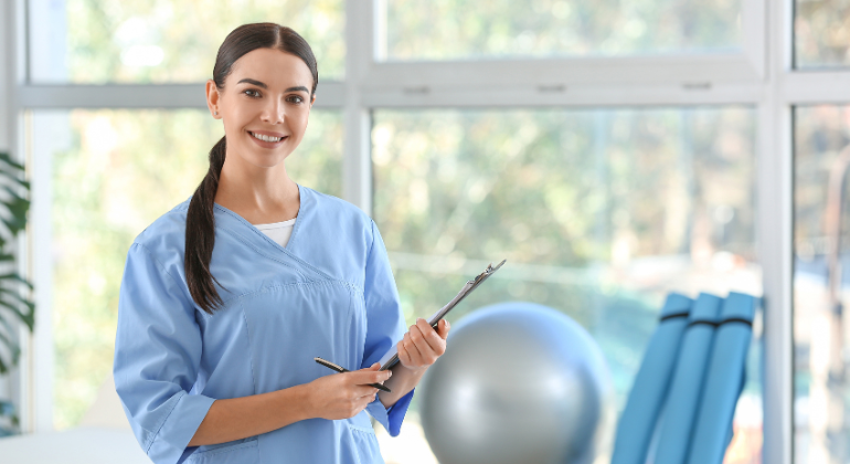 Smiling female physiotherapist in a light blue uniform holding a clipboard, standing in a bright, well-equipped therapy room with exercise equipment in the background.