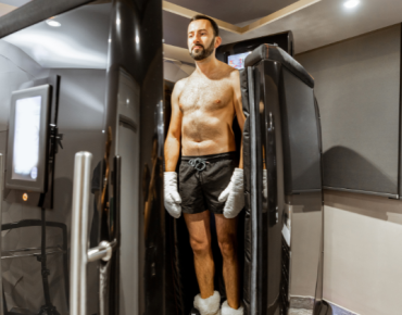 A man is standing inside a cryotherapy chamber. He is shirtless, wearing black shorts, white gloves, and socks, with his eyes closed, appearing calm and relaxed as the treatment begins. The chamber is sleek and modern, with a digital screen on the side displaying settings and controls. The room has a minimalist design with neutral tones, enhancing the clinical yet comfortable atmosphere.