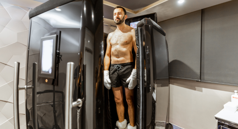 A man is standing inside a cryotherapy chamber. He is shirtless, wearing black shorts, white gloves, and socks, with his eyes closed, appearing calm and relaxed as the treatment begins. The chamber is sleek and modern, with a digital screen on the side displaying settings and controls. The room has a minimalist design with neutral tones, enhancing the clinical yet comfortable atmosphere.