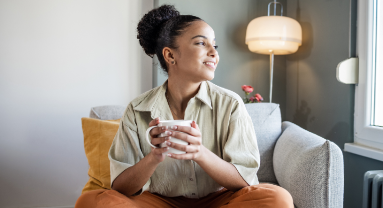 A woman is sitting comfortably on a couch, holding a white mug with both hands and smiling as she looks out the window. She has her hair styled in a neat bun and is dressed in a light-colored blouse and orange pants. The room is cozy, featuring soft lighting from a modern lamp and neutral-colored decor, creating a warm and relaxed atmosphere. A yellow cushion is placed behind her, adding a pop of color to the setting.