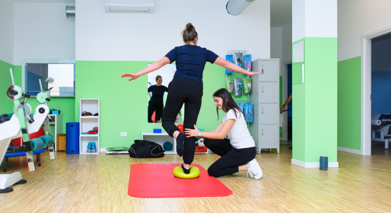 The image shows a physiotherapy session taking place in a brightly lit, well-equipped clinic. A patient is balancing on one foot on a green balance cushion while a physiotherapist supports and guides her. The patient is standing on a red exercise mat, arms extended for balance, with the physiotherapist kneeling beside her, ensuring proper posture and technique. The room features modern rehabilitation equipment and green walls, creating a calm and focused environment for therapy.