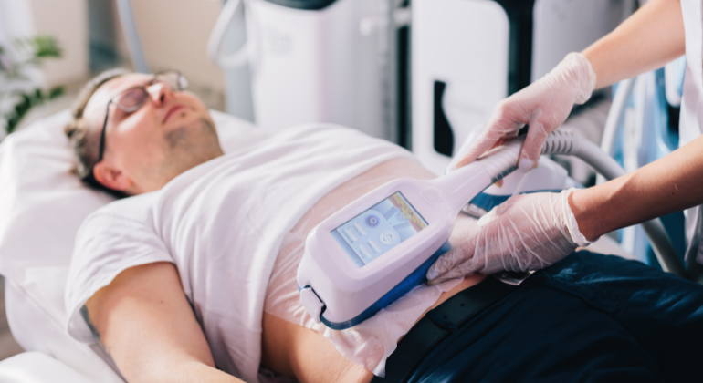 A man is lying down on a treatment bed, receiving a cryolipolysis (fat-freezing) procedure on his abdomen. He is wearing glasses and a white shirt, appearing relaxed with his eyes closed. A healthcare professional, wearing gloves, is holding a cryolipolysis device that is attached to the man's abdomen. The device's screen displays treatment settings. The clinical environment is clean and modern, with medical equipment visible in the background.