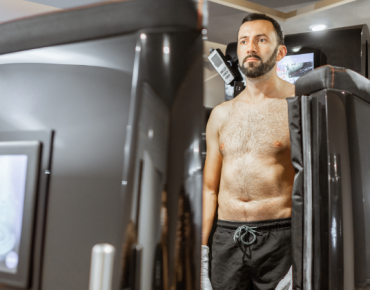 The image shows a man undergoing a whole-body cryotherapy session. He is standing inside a cryotherapy chamber, with the top half of his body exposed while the lower half is enclosed within the chamber. The man is wearing black shorts and appears calm and focused as he experiences the extreme cold temperatures used in cryotherapy. The chamber is sleek and modern, equipped with control panels and displays, highlighting the advanced technology used in cryotherapy treatments. This image captures the process of cryotherapy, emphasizing its role in wellness and recovery therapies.