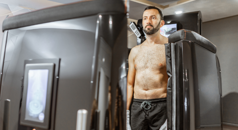 The image shows a man undergoing a whole-body cryotherapy session. He is standing inside a cryotherapy chamber, with the top half of his body exposed while the lower half is enclosed within the chamber. The man is wearing black shorts and appears calm and focused as he experiences the extreme cold temperatures used in cryotherapy. The chamber is sleek and modern, equipped with control panels and displays, highlighting the advanced technology used in cryotherapy treatments. This image captures the process of cryotherapy, emphasizing its role in wellness and recovery therapies.