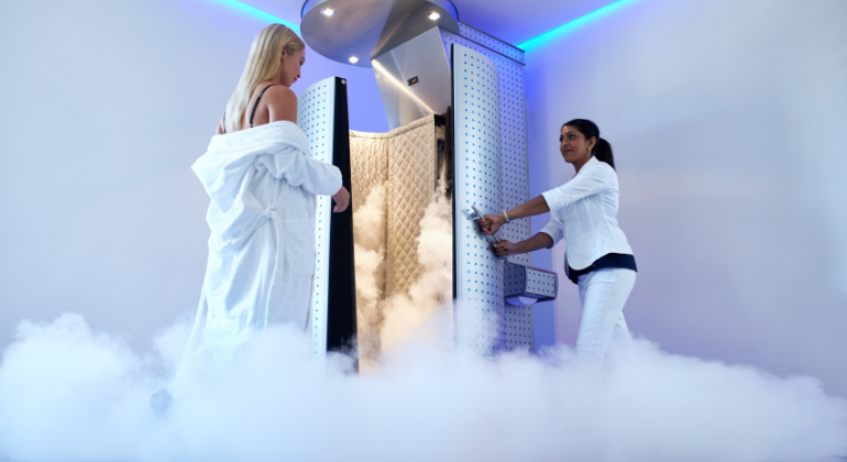 Therapist opening a cryotherapy chamber door as a woman in a robe prepares for her whole-body cryotherapy session surrounded by cold mist.