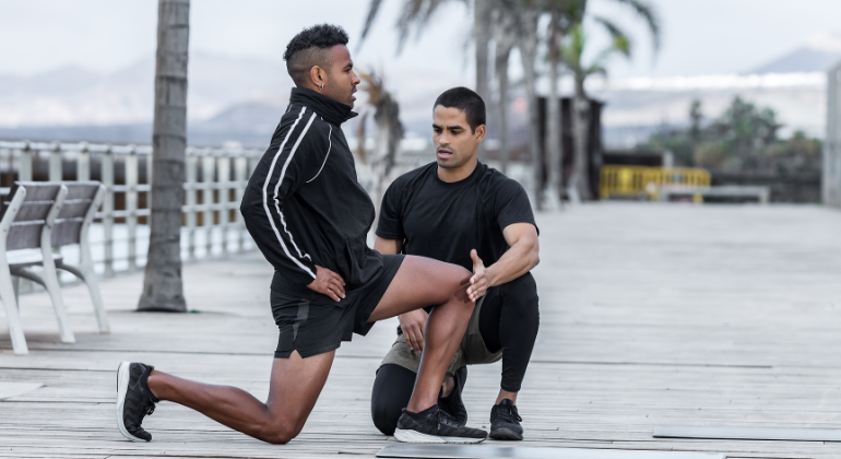 The image shows an outdoor physiotherapy or training session taking place on a boardwalk with a scenic backdrop of palm trees and mountains. A trainer or physiotherapist is assisting a male athlete with a deep lunge stretch, focusing on proper form and technique. The athlete, dressed in black sportswear, is stretching one leg forward while keeping the other knee on the ground, working on flexibility and strength. The trainer is closely monitoring and supporting the athlete’s movement, ensuring the exercise is performed correctly to prevent injury. The setting provides a refreshing and motivating environment for physical therapy or athletic training.