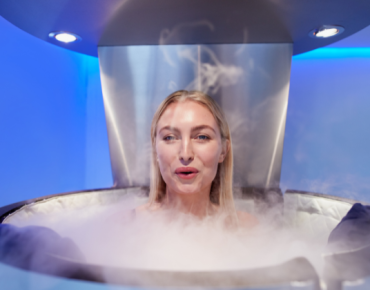 Person undergoing a cryotherapy session inside a chamber, with cold mist surrounding them in a blue-lit room.