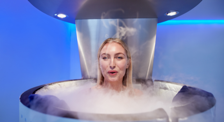 Person undergoing a cryotherapy session inside a chamber, with cold mist surrounding them in a blue-lit room.