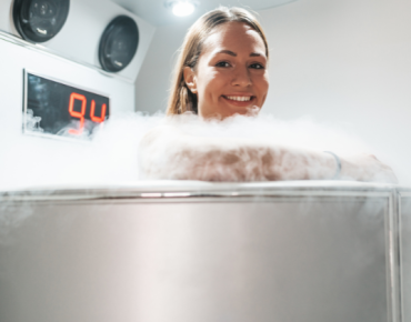 Smiling woman undergoing whole-body cryotherapy session in a cryotherapy chamber surrounded by cold mist.