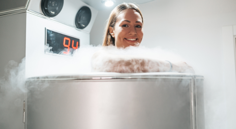 Smiling woman undergoing whole-body cryotherapy session in a cryotherapy chamber surrounded by cold mist.