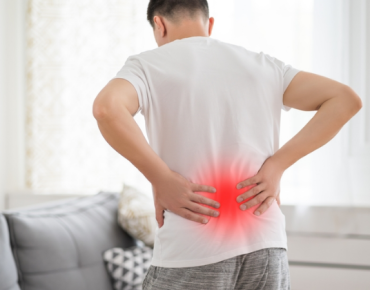 Man holding his lower back in pain, with a red highlighted area to indicate the location of discomfort, standing in a home environment. The image symbolizes chronic pain, possibly related to conditions like back pain or muscle strain.