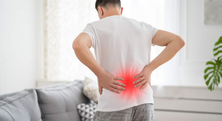 Man holding his lower back in pain, with a red highlighted area to indicate the location of discomfort, standing in a home environment. The image symbolizes chronic pain, possibly related to conditions like back pain or muscle strain.