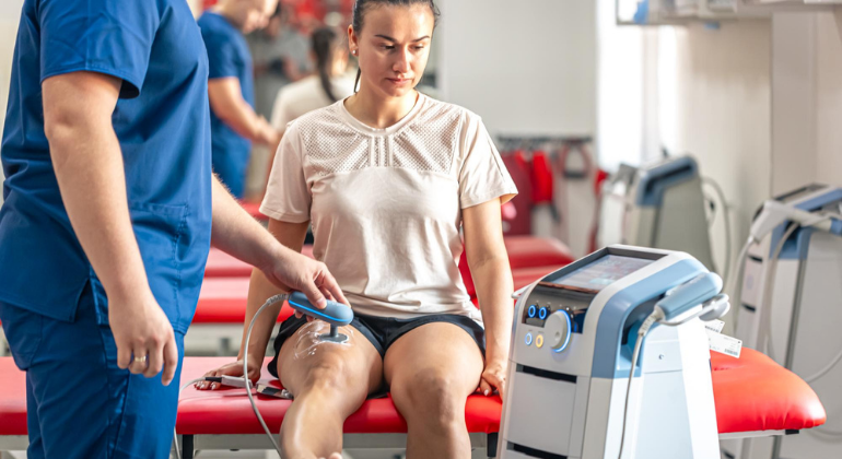 A physiotherapist performs localized cryotherapy on a female athlete's knee using a handheld device. The athlete sits on a treatment table in a clinical setting, receiving cold therapy to reduce pain and inflammation in the joint area.