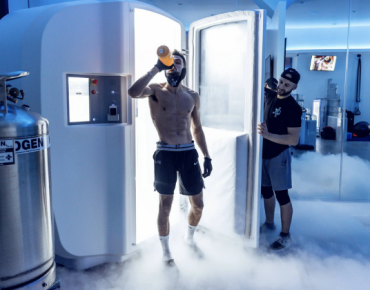 An athlete steps out of a cryotherapy chamber surrounded by cold vapor, wearing protective gloves and drinking from a bottle post-session. A technician assists nearby, with nitrogen tanks visible in the background used to cool the chamber for whole-body cryotherapy.