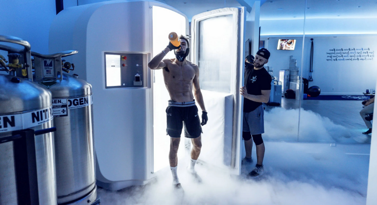 An athlete steps out of a cryotherapy chamber surrounded by cold vapor, wearing protective gloves and drinking from a bottle post-session. A technician assists nearby, with nitrogen tanks visible in the background used to cool the chamber for whole-body cryotherapy.