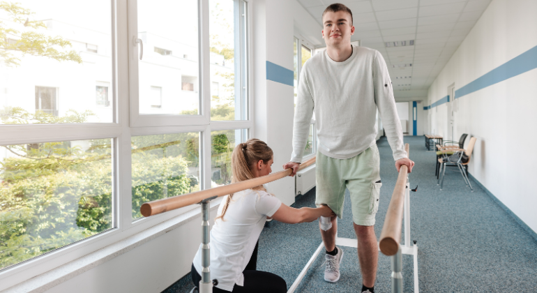 Physiotherapist assisting a young male patient with walking rehabilitation using parallel bars during a recovery session.