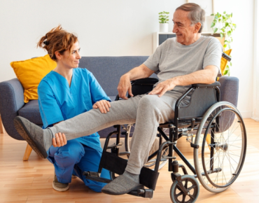 Physiotherapist helping an elderly man in a wheelchair with leg mobility exercises during a home physiotherapy session in a warm, well-lit living space.