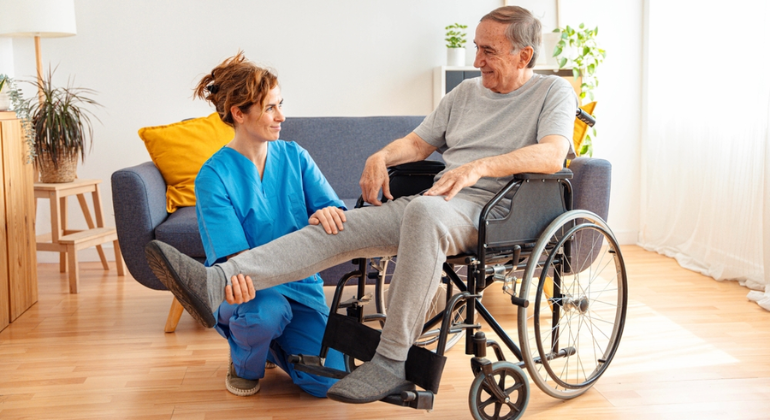 Physiotherapist helping an elderly man in a wheelchair with leg mobility exercises during a home physiotherapy session in a warm, well-lit living space.