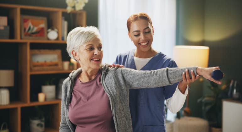 Physiotherapist guiding an elderly woman through arm strengthening exercises with a dumbbell during a home physiotherapy session in a comfortable setting.