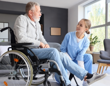 Physiotherapist assisting an elderly man in a wheelchair with leg exercises during a home physiotherapy session in a bright, modern living room.