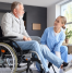 Physiotherapist assisting an elderly man in a wheelchair with leg exercises during a home physiotherapy session in a bright, modern living room.