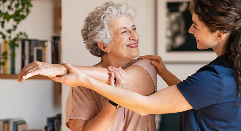 Physiotherapist assisting an elderly woman with arm and shoulder stretching exercises during a home physiotherapy session in a warm and friendly environment.