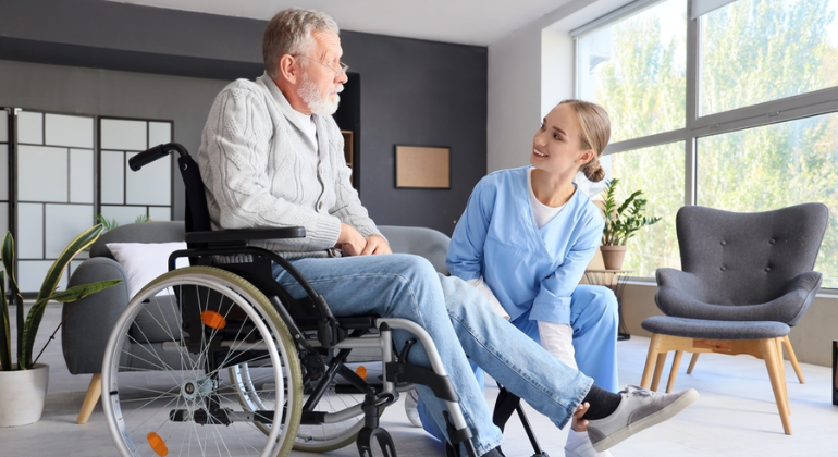 Physiotherapist assisting an elderly man in a wheelchair with leg exercises during a home physiotherapy session in a bright, modern living room.