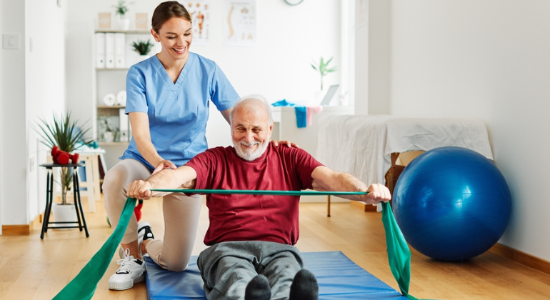 Physiotherapist guiding an elderly man through resistance band exercises during a home physiotherapy session in a bright and organized therapy space.