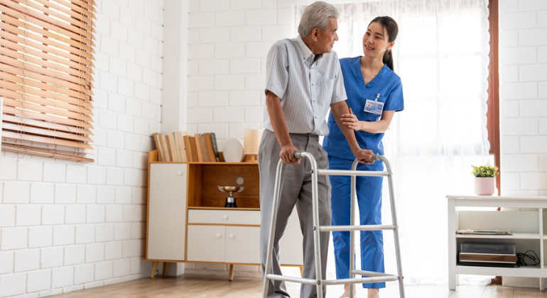 Physiotherapist supporting an elderly man using a walker during a home physiotherapy session in a bright and organized room.