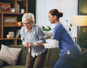 Physiotherapist assisting an elderly woman with a walking stick during a home physiotherapy session in a comfortable living room setting.