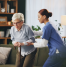 Physiotherapist assisting an elderly woman with a walking stick during a home physiotherapy session in a comfortable living room setting.