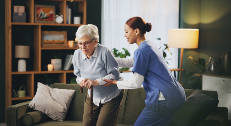 Physiotherapist assisting an elderly woman with a walking stick during a home physiotherapy session in a comfortable living room setting.