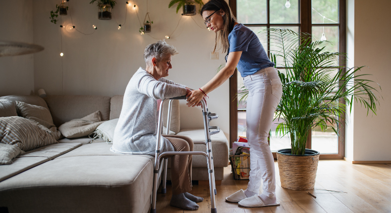 Physiotherapist supporting an elderly woman using a walker during a home physiotherapy session in a cozy living room with natural light and greenery.